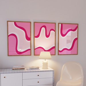 May include: Three framed abstract art prints with pink and white wavy lines on a white wall above a white dresser with three drawers and a lamp on top. A white chair is in the foreground.