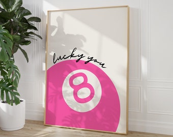 May include: A pink and white 8-ball with the text "lucky you" in black script. The 8-ball is in a gold frame and is hanging on a white wall.