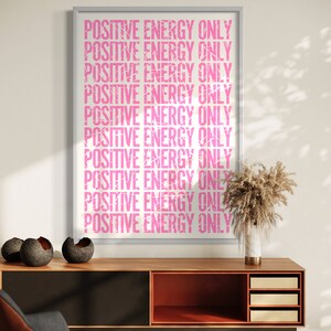Może przedstawiać: Wydruk w ramie z tekstem "POSITIVE ENERGY ONLY" powt&oacute;rzonym r&oacute;żowymi literami na białym tle. Wydruk wisi na białej ścianie nad drewnianą szafką z czarnym i pomarańczowym wnętrzem.