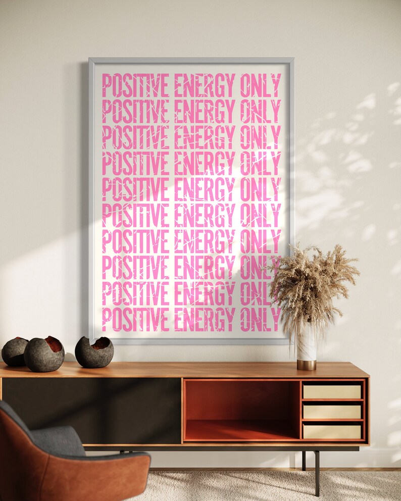 Może przedstawiać: Wydruk w ramie z tekstem "POSITIVE ENERGY ONLY" powt&oacute;rzonym r&oacute;żowymi literami na białym tle. Wydruk wisi na białej ścianie nad drewnianą szafką z czarnym i pomarańczowym wnętrzem.