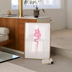 Pode incluir: Uma impress&atilde;o emoldurada de uma cobra rosa sobre um fundo branco. A cobra est&aacute; enrolada em forma de espiral e est&aacute; em uma sala com uma janela e uma l&acirc;mpada.