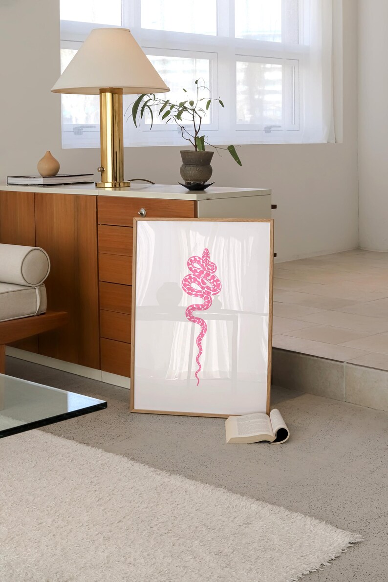 Pode incluir: Uma impress&atilde;o emoldurada de uma cobra rosa sobre um fundo branco. A cobra est&aacute; enrolada em forma de espiral e est&aacute; em uma sala com uma janela e uma l&acirc;mpada.