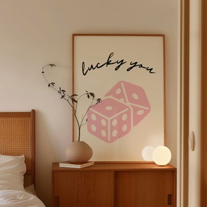 Pu&ograve; includere: Un muro bianco e beige con un quadro incorniciato in legno appeso ad esso. Il quadro presenta un disegno di dadi rosa e bianchi con il testo "lucky you" in nero. Un vaso marrone con un ramo frondoso &egrave; su un tavolo di legno con una lampada bianca. Il tavolo &egrave; davanti a un letto con una testiera bianca e biancheria da letto bianca.