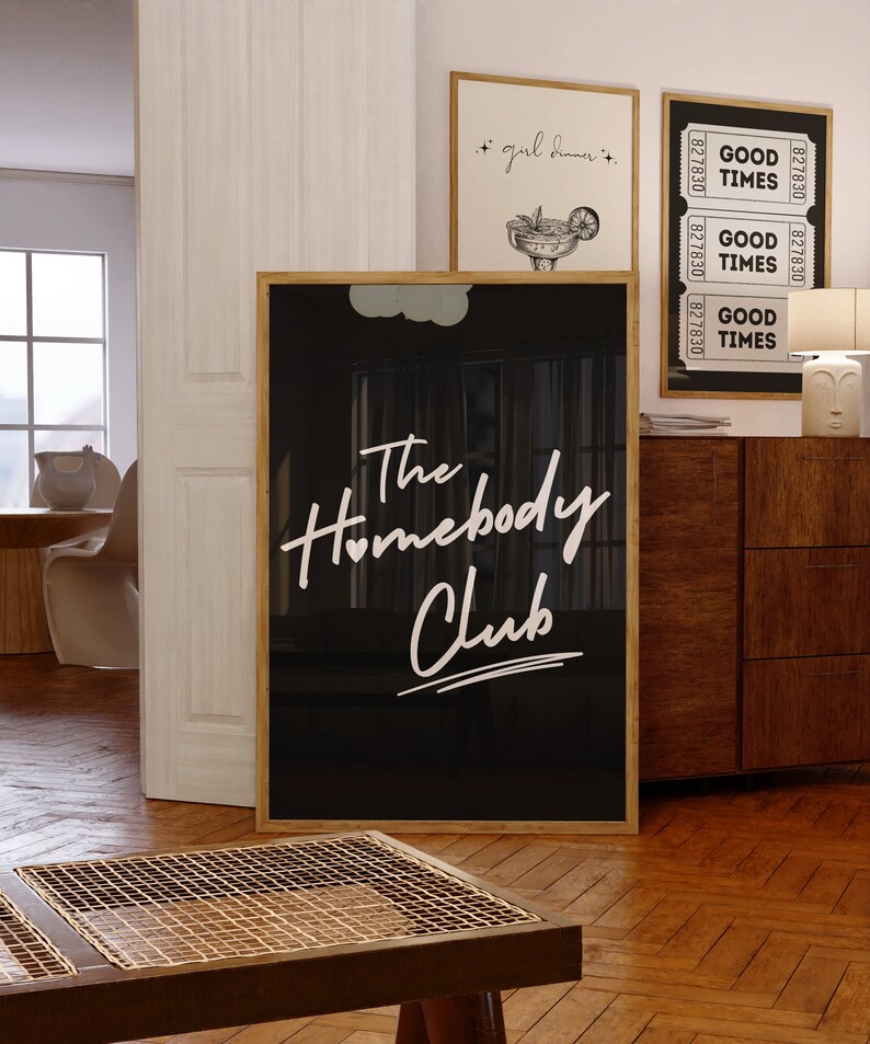 May include: Black and white framed print with the text "The Homebody Club" in white script font. The print is hanging on a wall in a room with a wooden floor.
