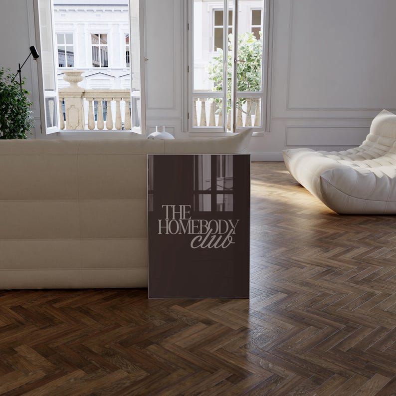May include: A brown rectangular sign with the text "THE HOMEBODY CLUB" in white. The sign is in a room with a white sofa, a white chair, and large windows overlooking a city.