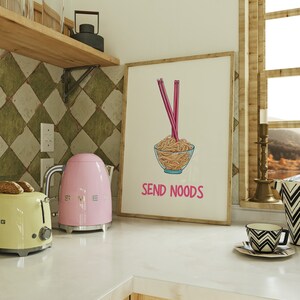 May include: A framed print with a cartoon illustration of noodles in a glass bowl with pink chopsticks. The text "SEND NOODS" is below the bowl. A pink kettle and yellow toaster are on the counter.