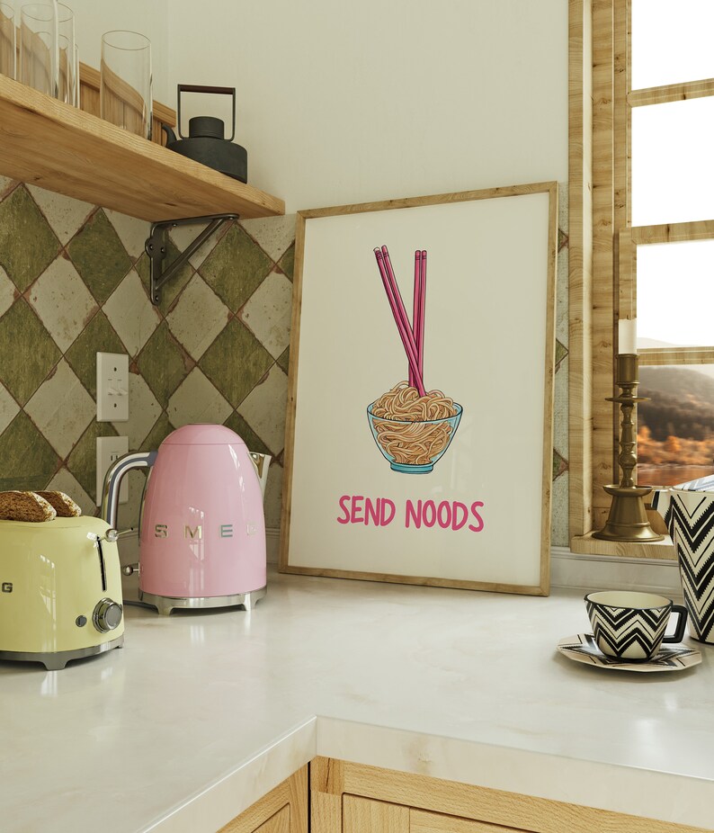 May include: A framed print with a cartoon illustration of noodles in a glass bowl with pink chopsticks. The text "SEND NOODS" is below the bowl. A pink kettle and yellow toaster are on the counter.