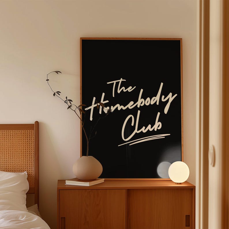 May include: A black framed print with white text that reads "The Homebody Club" hanging on a wall above a wooden dresser with a white lamp and a vase with a plant.