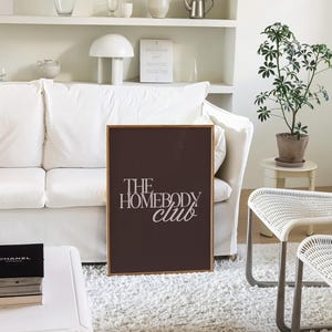 May include: A framed print with the text "THE HOMEBODY club" in white script on a dark brown background. The print is displayed in a living room setting with a white sofa, rug, and decor.