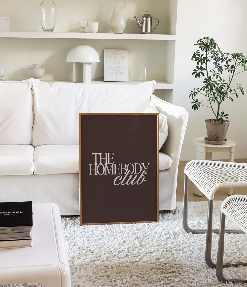May include: A framed print with the text "THE HOMEBODY club" in white script on a dark brown background. The print is displayed in a living room setting with a white sofa, rug, and decor.