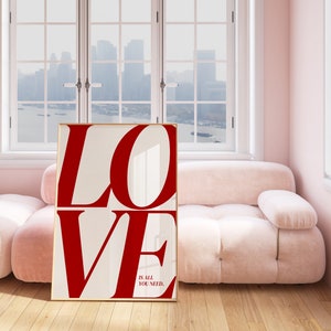 May include: A white framed poster with the word "LOVE" in large red letters. The text "IS ALL YOU NEED." is printed in smaller black letters below the word "LOVE". The poster is leaning against a pink sofa in a room with a window.