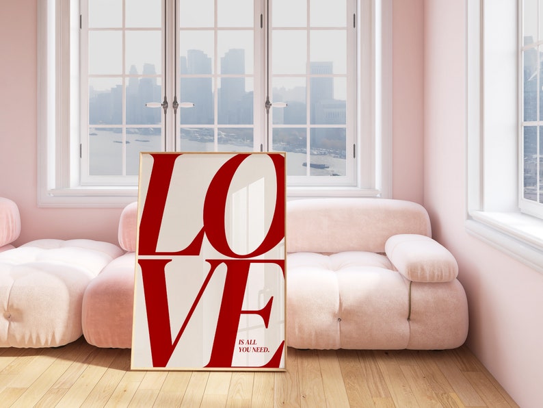 May include: A white framed poster with the word "LOVE" in large red letters. The text "IS ALL YOU NEED." is printed in smaller black letters below the word "LOVE". The poster is leaning against a pink sofa in a room with a window.
