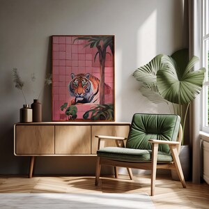 May include: A framed print of a tiger peering out from behind a pink tiled wall with green plants. The print is hanging above a wooden cabinet and a green armchair in a modern living room.