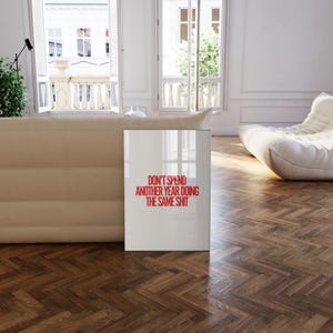 May include: A white poster with red text that reads "Don't spend another year doing the same shit." The poster is leaning against a white couch in a room with a white floor and white walls.