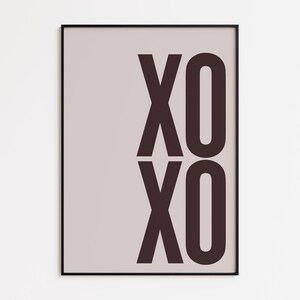 May include: A framed art print featuring the letters "XO" stacked vertically in a dark brown color. The background is a light beige, and the frame is black. The artwork is a simple, modern design.