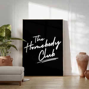 May include: A black framed poster with white text that reads "The Homebody Club" with a small heart symbol in the word "Homebody". The poster is hanging on a white wall in a room with a potted plant, a light gray ottoman, and two terracotta pots.
