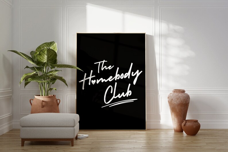 May include: A black framed poster with white text that reads "The Homebody Club" with a small heart symbol in the word "Homebody". The poster is hanging on a white wall in a room with a potted plant, a light gray ottoman, and two terracotta pots.
