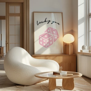 Pu&ograve; includere: Una poltrona bianca e soffice con un tavolino da caff&egrave; in legno di fronte. Il tavolino da caff&egrave; ha un piano in vetro e una base in legno. Una stampa incorniciata con il testo "lucky you" e un design a dadi rosa &egrave; appesa al muro dietro la poltrona. Una lampada bianca con un paralume rotondo si trova su uno scaffale in legno.