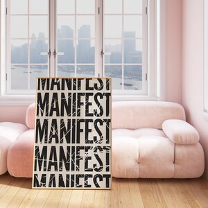 Pode incluir: Um p&ocirc;ster emoldurado com a palavra "MANIFEST" repetida cinco vezes em texto preto sobre fundo branco. O p&ocirc;ster est&aacute; encostado em um sof&aacute; rosa claro em uma sala com grandes janelas.