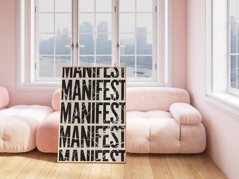Pode incluir: Um p&ocirc;ster emoldurado com a palavra "MANIFEST" repetida cinco vezes em texto preto sobre fundo branco. O p&ocirc;ster est&aacute; encostado em um sof&aacute; rosa claro em uma sala com grandes janelas.