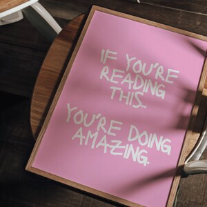 May include: A pink framed sign with white lettering that reads "If you're reading this... you're doing amazing."