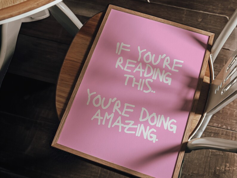 May include: A pink framed sign with white lettering that reads "If you're reading this... you're doing amazing."