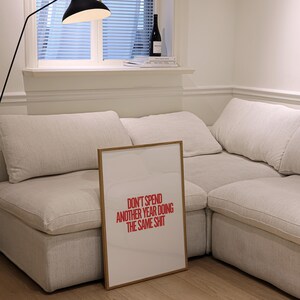 May include: A framed print with a white background and red text that reads "Don't spend another year doing the same shit". The print is leaning against a white couch in a living room.