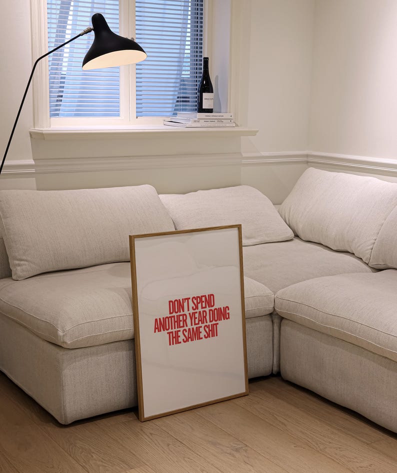 May include: A framed print with a white background and red text that reads "Don't spend another year doing the same shit". The print is leaning against a white couch in a living room.