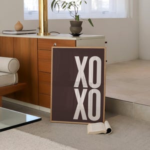 Może przedstawiać: Oprawiony wydruk z napisem "XO XO" w kolorze białym na ciemnobrązowym tle. Wydruk opiera się o ścianę w pokoju z lampą, małą rośliną i książką. Pok&oacute;j ma neutralną paletę kolor&oacute;w.