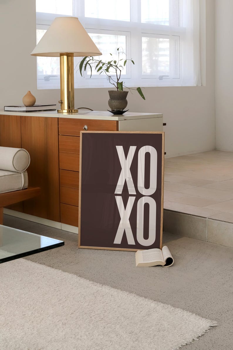Może przedstawiać: Oprawiony wydruk z napisem "XO XO" w kolorze białym na ciemnobrązowym tle. Wydruk opiera się o ścianę w pokoju z lampą, małą rośliną i książką. Pok&oacute;j ma neutralną paletę kolor&oacute;w.