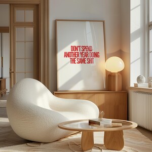 May include: A white, textured armchair with a round, wooden coffee table in front of it. A framed poster with the text "Don't spend another year doing the same shit" hangs on the wall behind the armchair. A wooden lamp with a white, round shade sits on a wooden shelf.