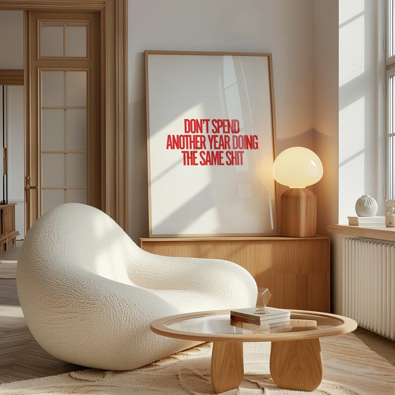 May include: A white, textured armchair with a round, wooden coffee table in front of it. A framed poster with the text "Don't spend another year doing the same shit" hangs on the wall behind the armchair. A wooden lamp with a white, round shade sits on a wooden shelf.