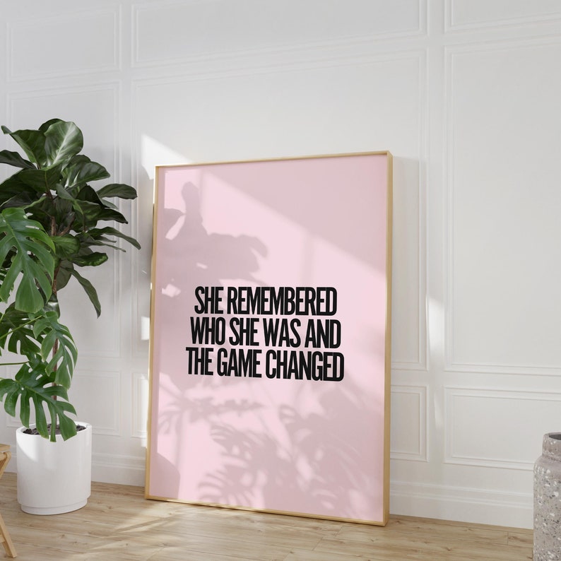 K&ouml;nnte beinhalten: Ein gerahmter rosa Kunstdruck mit dem Text "SHE REMEMBERED WHO SHE WAS AND THE GAME CHANGED" in Schwarz. Der Rahmen hat eine helle Holzfarbe. Links vom Druck befindet sich eine gro&szlig;e gr&uuml;ne Pflanze in einem wei&szlig;en Topf.