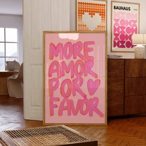 May include: Pink and orange poster with the text "MORE AMOR POR FAVOR" in a bold, handwritten style. The poster is framed in a light wood frame and is hanging on a wall in a room with a wooden floor.