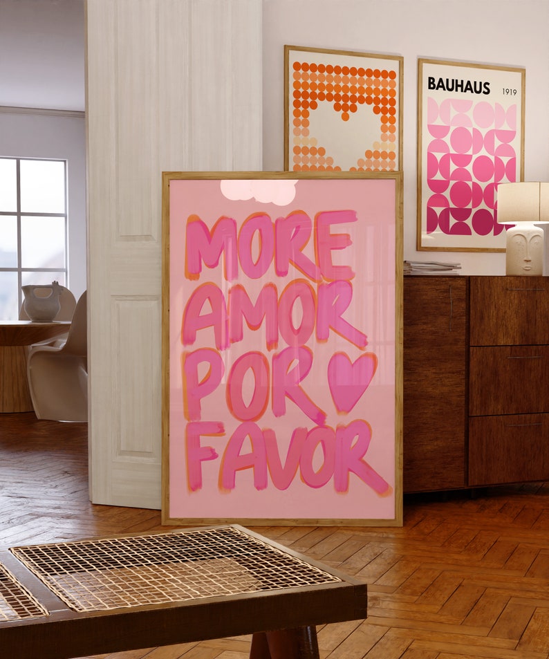 May include: Pink and orange poster with the text "MORE AMOR POR FAVOR" in a bold, handwritten style. The poster is framed in a light wood frame and is hanging on a wall in a room with a wooden floor.
