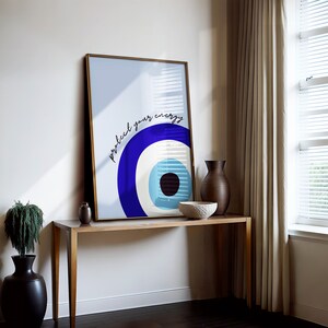 May include: A framed print with a blue and white evil eye design and the text "protect your energy". The print is hanging on a white wall above a wooden table.