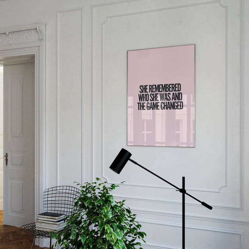 K&ouml;nnte beinhalten: Ein gerahmter rosa Druck mit dem schwarzen Text "SHE REMEMBERED WHO SHE WAS AND THE GAME CHANGED". Der Druck h&auml;ngt an einer wei&szlig;en Wand. Eine schwarze Lampe und eine gr&uuml;ne Pflanze befinden sich im Vordergrund.