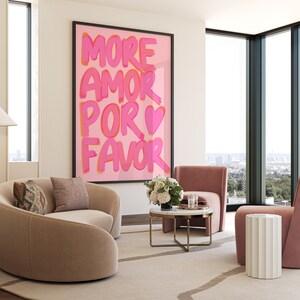 May include: A living room interior with a pink and white framed print that reads "MORE AMOR POR FAVOR" with a pink heart. The room features a light brown curved sofa, a white side table, a pink armchair, and a coffee table with a marble top and gold legs.