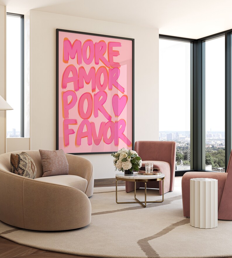 May include: A living room interior with a pink and white framed print that reads "MORE AMOR POR FAVOR" with a pink heart. The room features a light brown curved sofa, a white side table, a pink armchair, and a coffee table with a marble top and gold legs.
