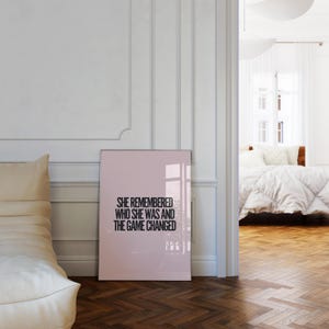 K&ouml;nnte beinhalten: Ein gerahmtes rosa Poster mit schwarzem Text, der lautet: "SHE REMEMBERED WHO SHE WAS AND THE GAME CHANGED", steht an einer wei&szlig;en Wand. Links ist ein beiger Sessel, und im Hintergrund ist ein Schlafzimmer zu sehen.