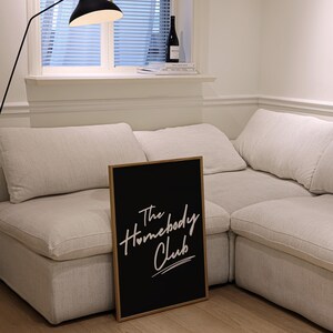 May include: A black framed sign with white text that reads "The Homebody Club" is leaning against a white sectional sofa. The sofa is in a room with a window and a floor lamp.