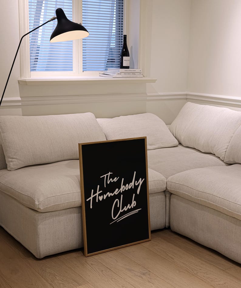 May include: A black framed sign with white text that reads "The Homebody Club" is leaning against a white sectional sofa. The sofa is in a room with a window and a floor lamp.