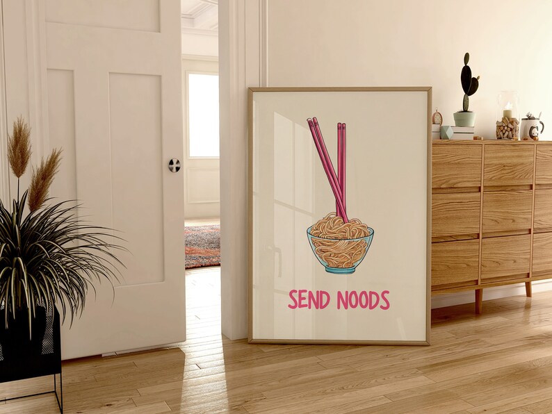 May include: A framed print featuring a bowl of noodles with pink chopsticks. The bowl is clear with a blue rim. The text "SEND NOODS" is below the bowl in pink. The print is displayed in a room with wooden floors.