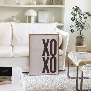May include: A framed art print with the text "XO XO" in dark brown letters. The print is displayed in a living room setting, leaning against a white sofa. The room features a white rug, a small plant, and white furniture.