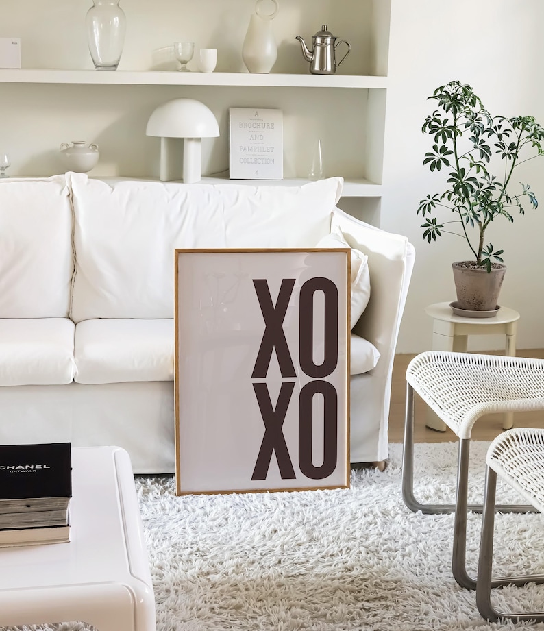 May include: A framed art print with the text "XO XO" in dark brown letters. The print is displayed in a living room setting, leaning against a white sofa. The room features a white rug, a small plant, and white furniture.