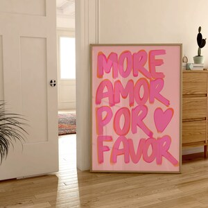 May include: Pink and orange text on a pink background reads "More Amor Por Favor" with a pink heart symbol. The print is framed and hanging on a wall.