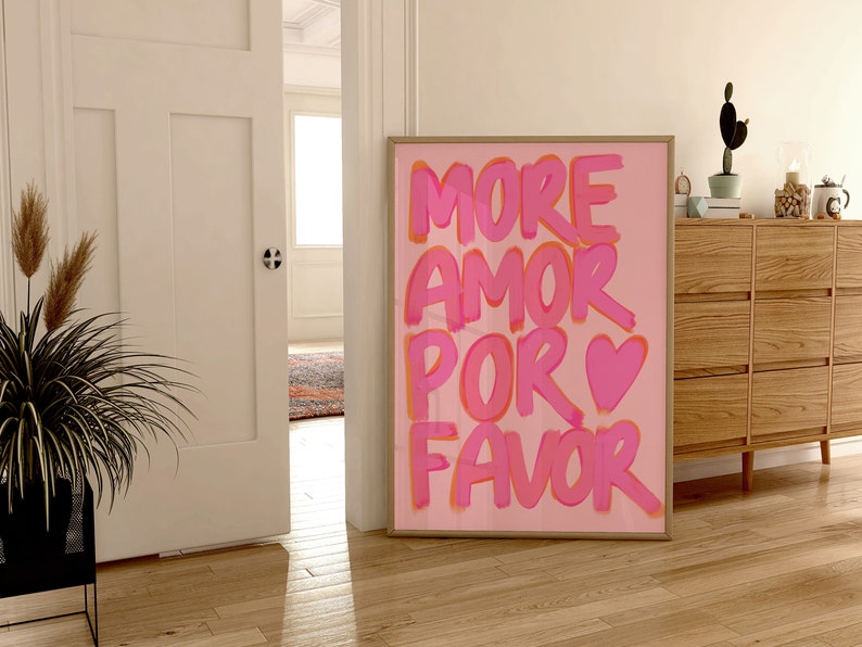 May include: Pink and orange text on a pink background reads "More Amor Por Favor" with a pink heart symbol. The print is framed and hanging on a wall.