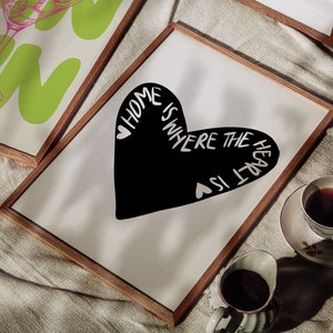 May include: A framed print with a black heart shape and the text "Home is where the heart is" in white. The print is on a white background and is framed in a natural wood frame.