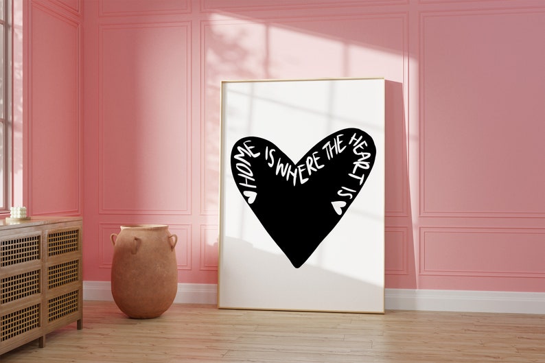 May include: A black and white print with a heart shape and the text "Home is where the heart is". The print is framed in a gold frame and is hanging on a pink wall.
