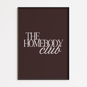 May include: A framed art print with the words "THE HOMEBODY club" in a stylish, light-colored font. The text is centered on a dark brown background, and the frame is black. The print is a decorative piece.